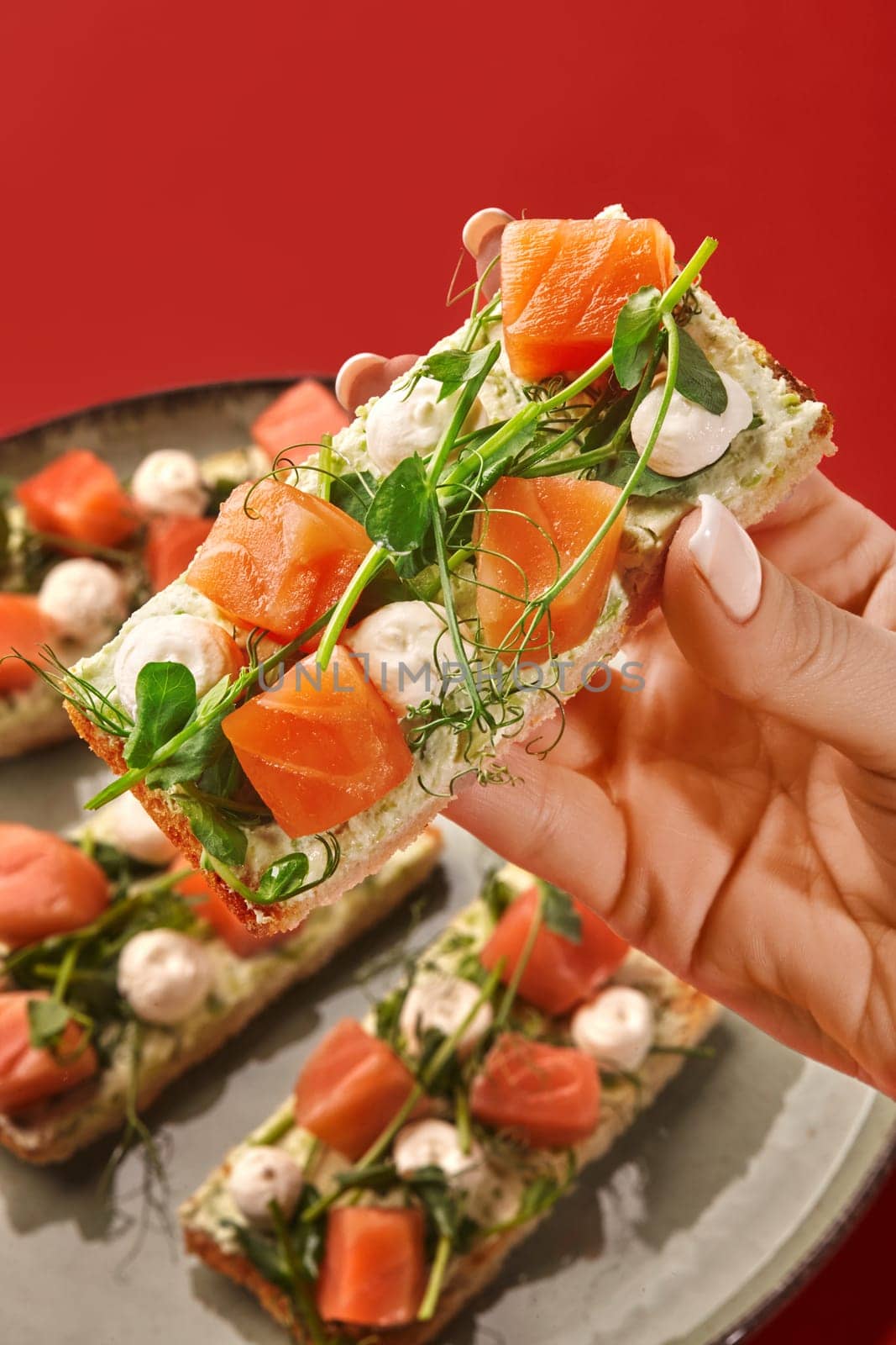 Female hand elegantly taking from plate appetizing crispy bruschetta topped with cream cheese, salmon gravlax, bocconcini and sprig of fresh herbs against red backdrop. Italian style snack