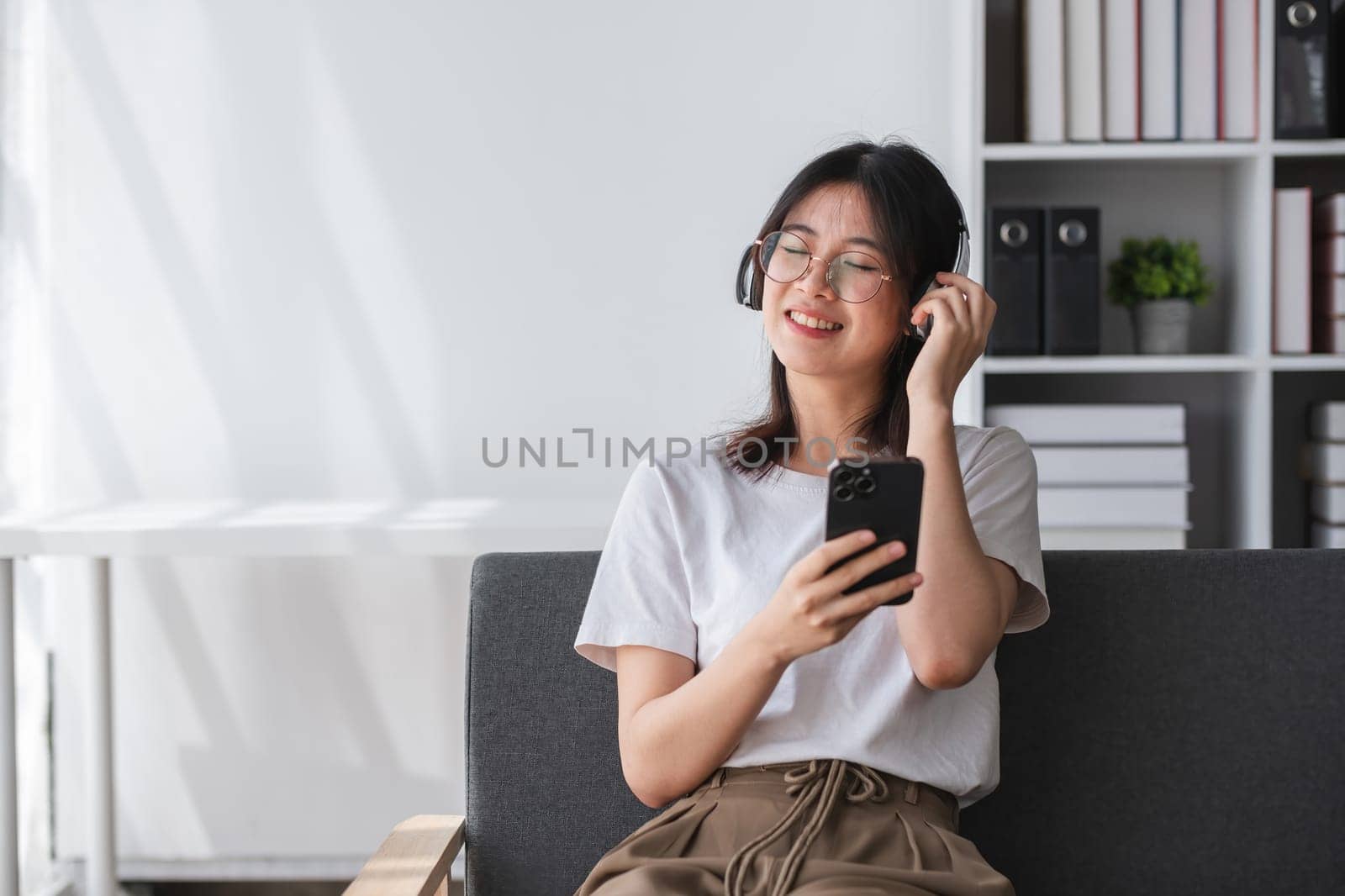 Happy Asian woman listening to music on mobile phone while sitting on sofa in living room on vacation..