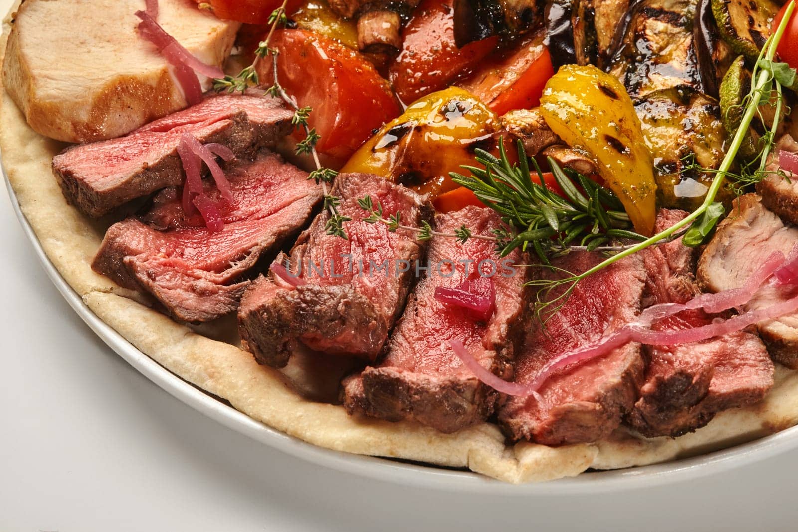 Closeup of sliced juicy roast beef with assorted grilled vegetables arranged on flatbread and garnished with aromatic herbs, on white background