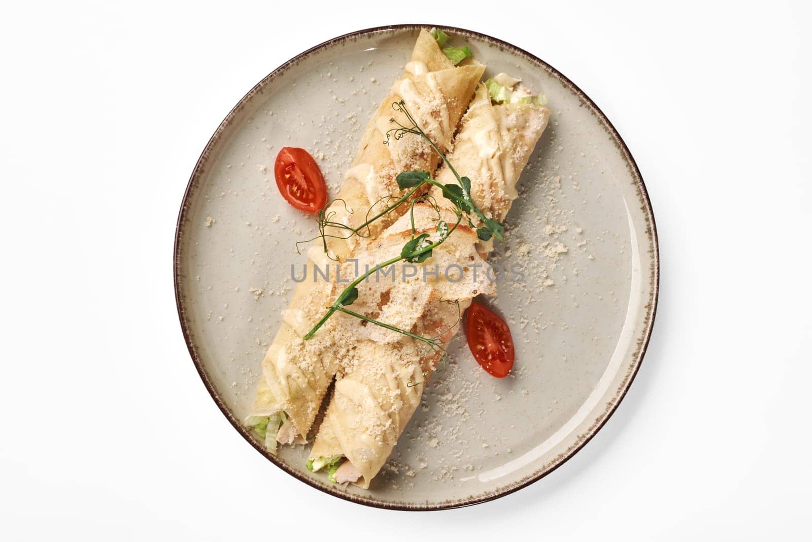 Top view of crepe rolls filled with lettuce and baked chicken, topped with rich Caesar dressing, crispy breadcrumbs and grated Parmesan, served with fresh tomato and greens on speckled plate