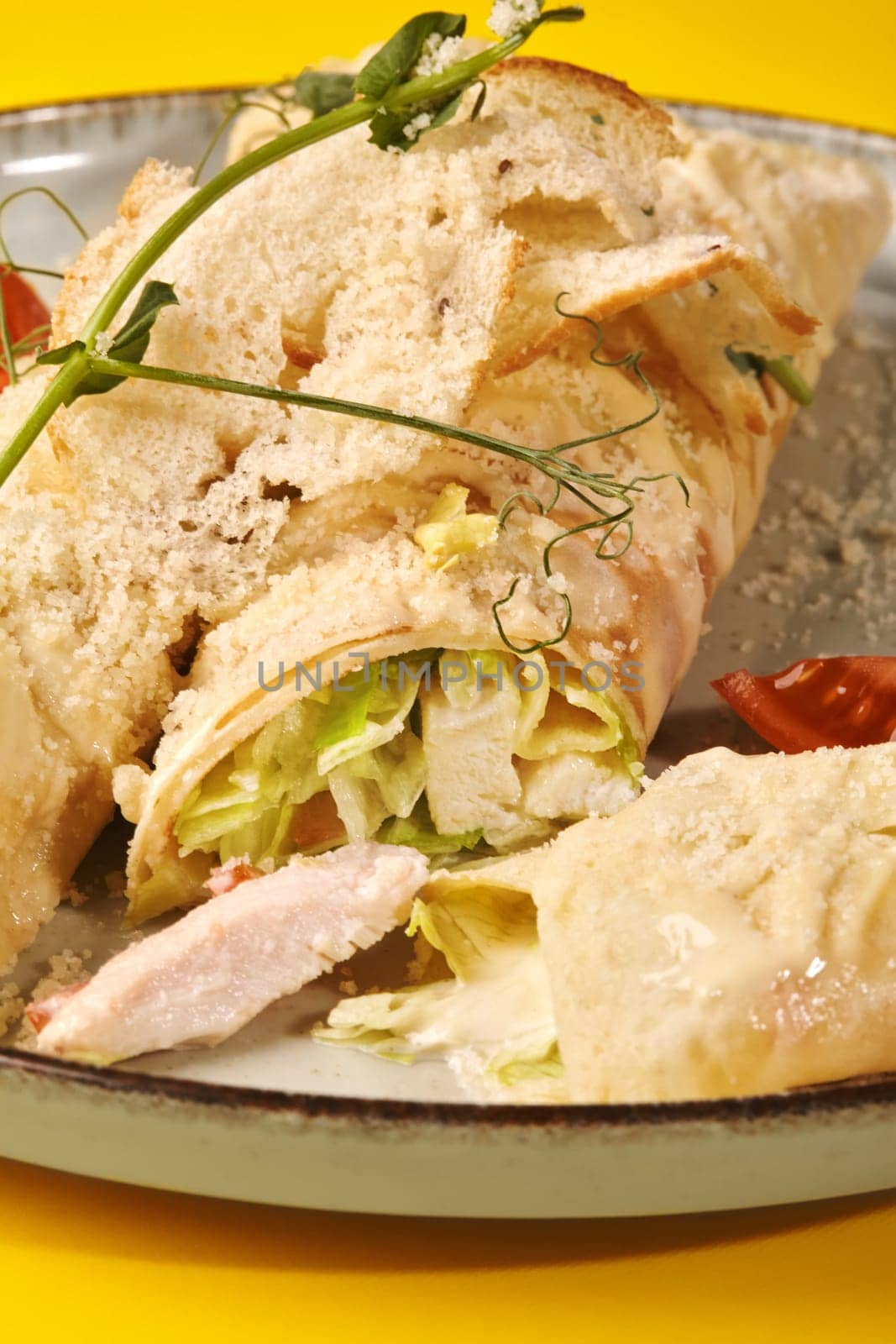 Closeup of delicious crepe rolls stuffed with lettuce and baked chicken, topped with creamy Caesar sauce, crispy breadcrumbs and grated Parmesan served on plate against yellow background