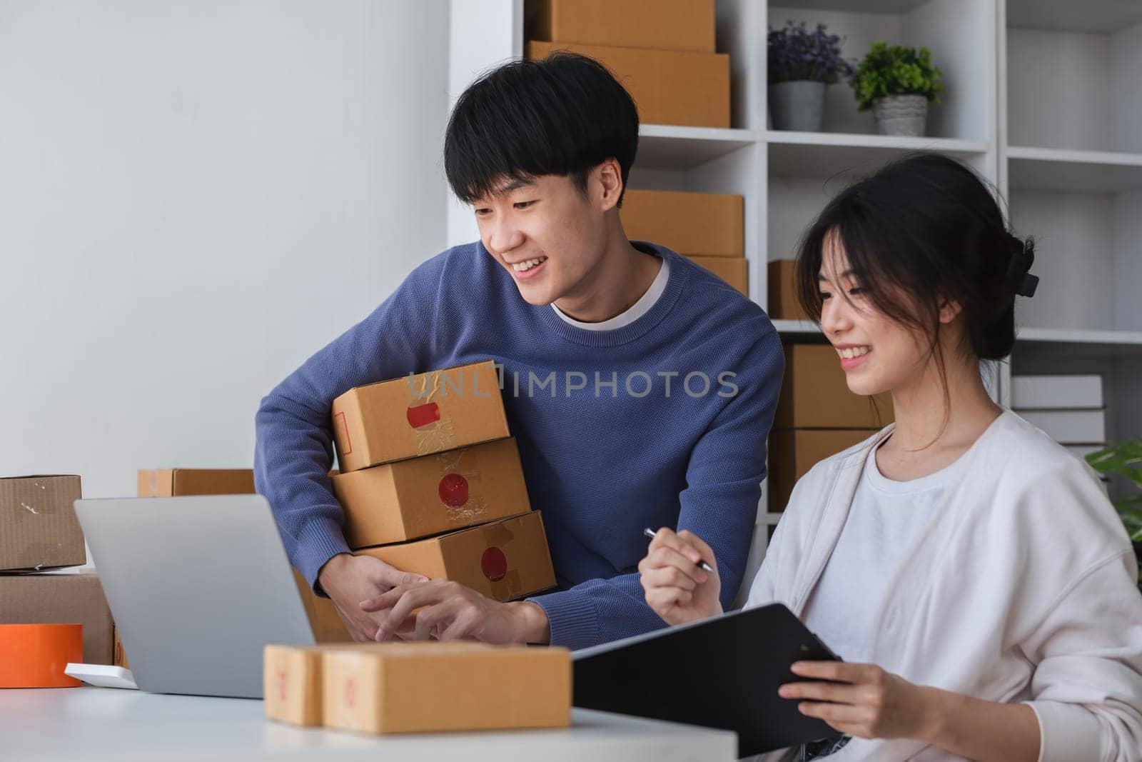 A happy Asian online shopping entrepreneur couple takes orders and packs products to send to customers together in their office. by wichayada