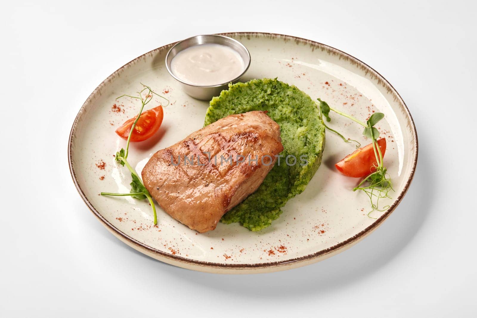 Healthy dinner of grilled chicken breast served with soft green potato puree with herbs and creamy dipping sauce garnished with cherry tomato and microgreens on ceramic plate