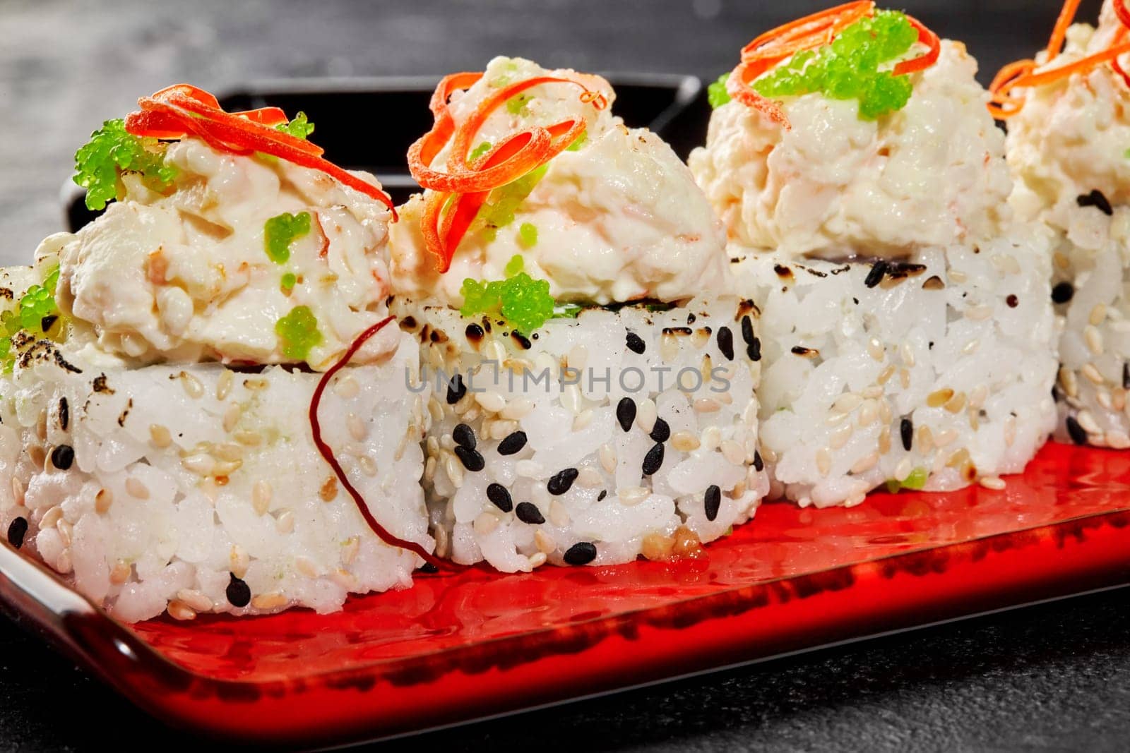 Macro shot of elegant sushi roll sprinkled with sesame seeds on ceramic red plate, highlighting creamy toppings with seafood garnished with green tobiko and vegetable shavings. Japanese style delicacy