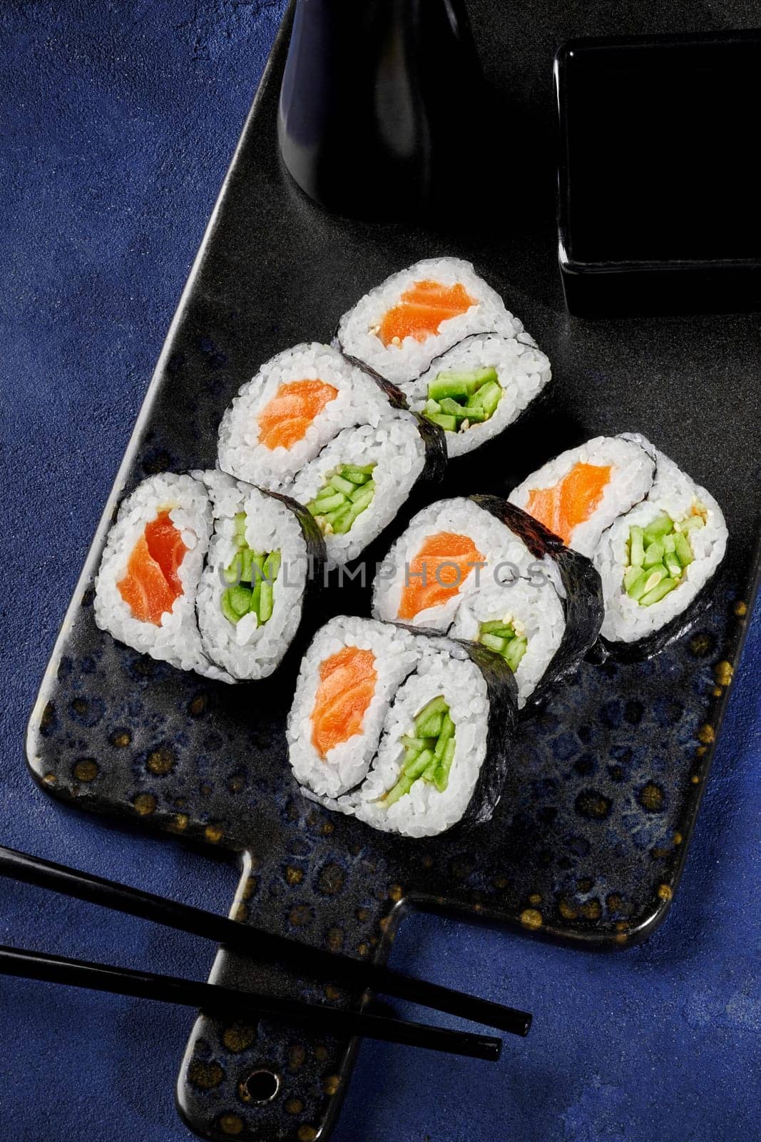 Delicious salmon and cucumber futomaki rolls with yin-yang design traditionally served with soy sauce on serving board against blue textured surface. Creative take on Japanese sushi presentation