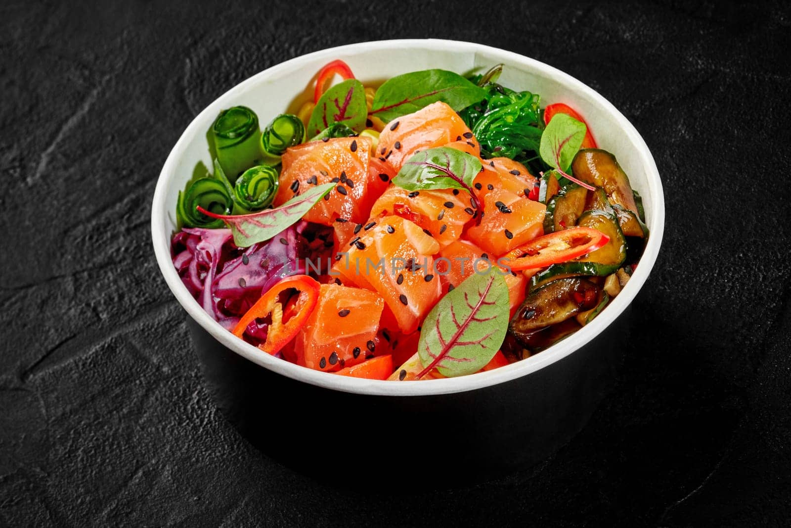 Vibrant salmon salad bursting with baked zucchini, fresh vegetables, greens and seaweed sprinkled with sesame seeds, served in cardboard bowl on dark background. Healthy snack. Delivery food