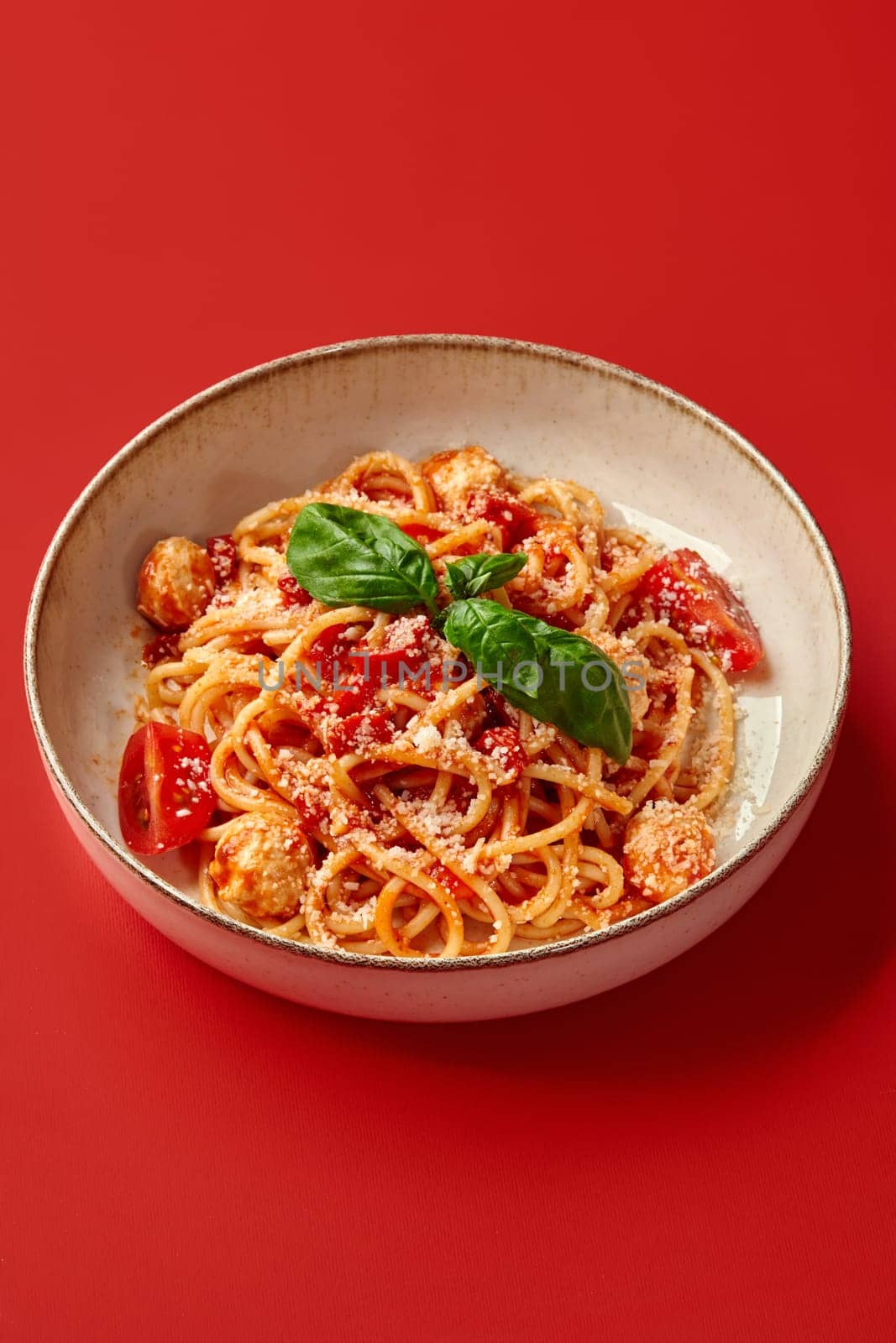 Appetizing spaghetti bowl with spicy tomato sauce and meatballs sprinkled with grated Parmesan cheese topped with fresh green basil leaves, served against vibrant red background
