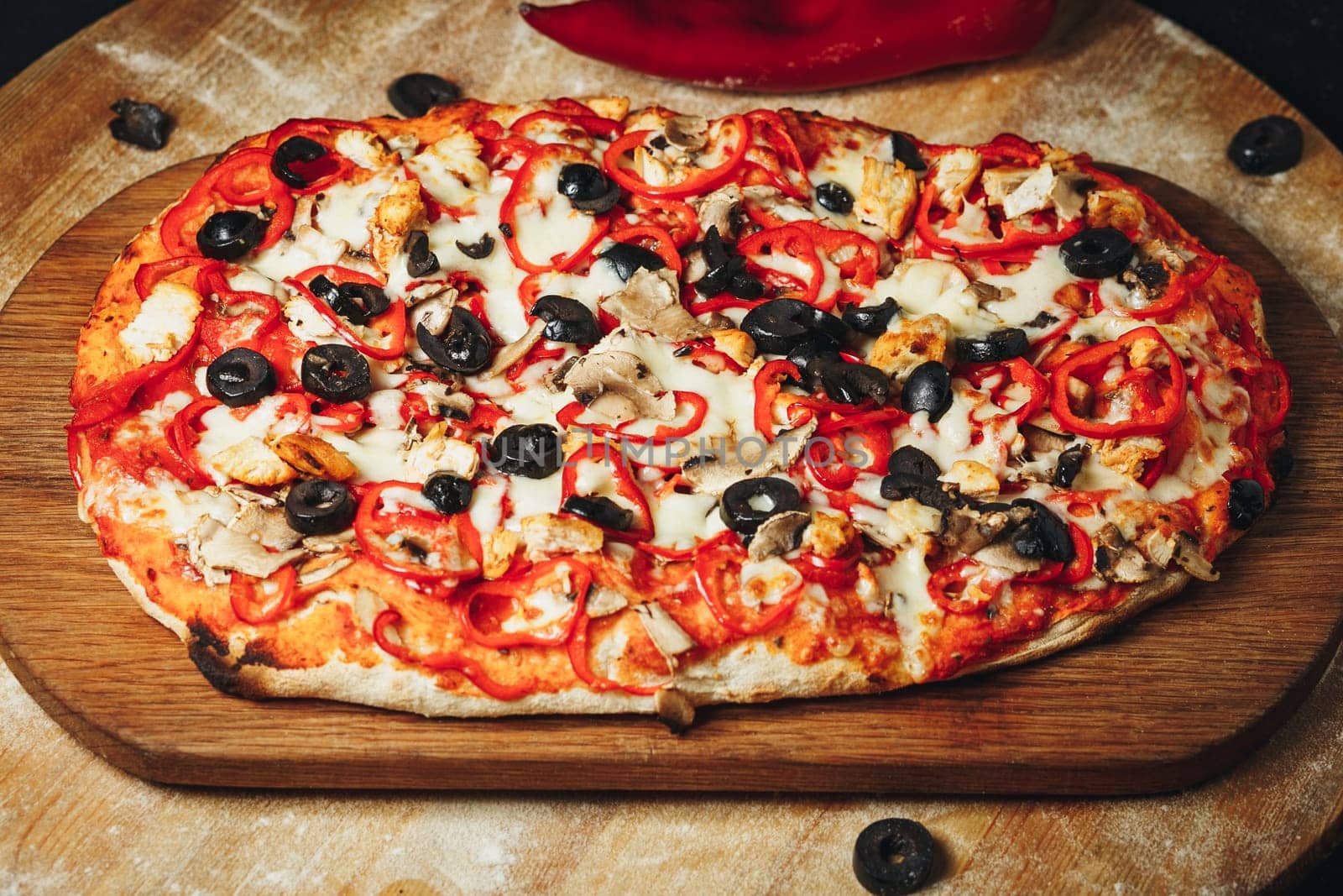 A freshly baked pizza topped with olives and peppers, resting on a rustic wooden cutting board.