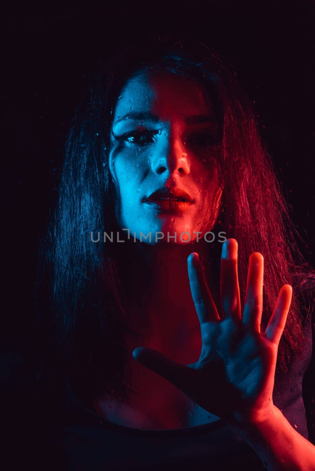 Sensual portrait of a beautiful attractive girl looking through the glass with raindrops with red blue lighting