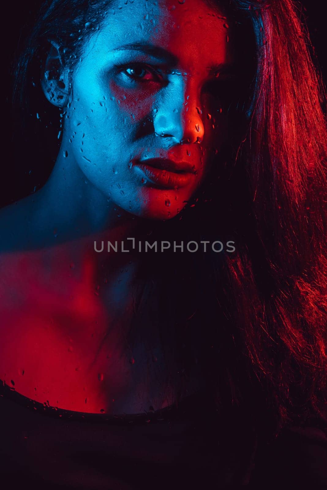 Sensual portrait of beautiful attractive brunette girl behind glass with raindrops with colored red blue neon lighting