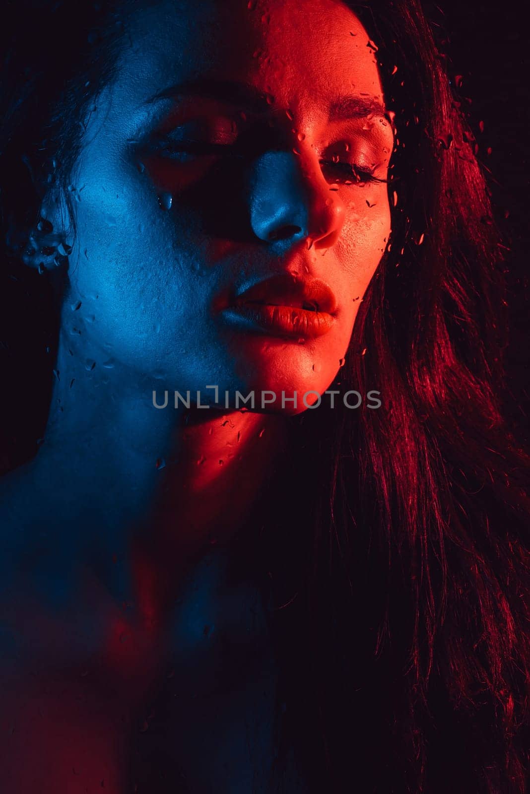 Sensual beauty fashion portrait of sexy attractive brunette girl through glass with raindrops and red blue neon lighting