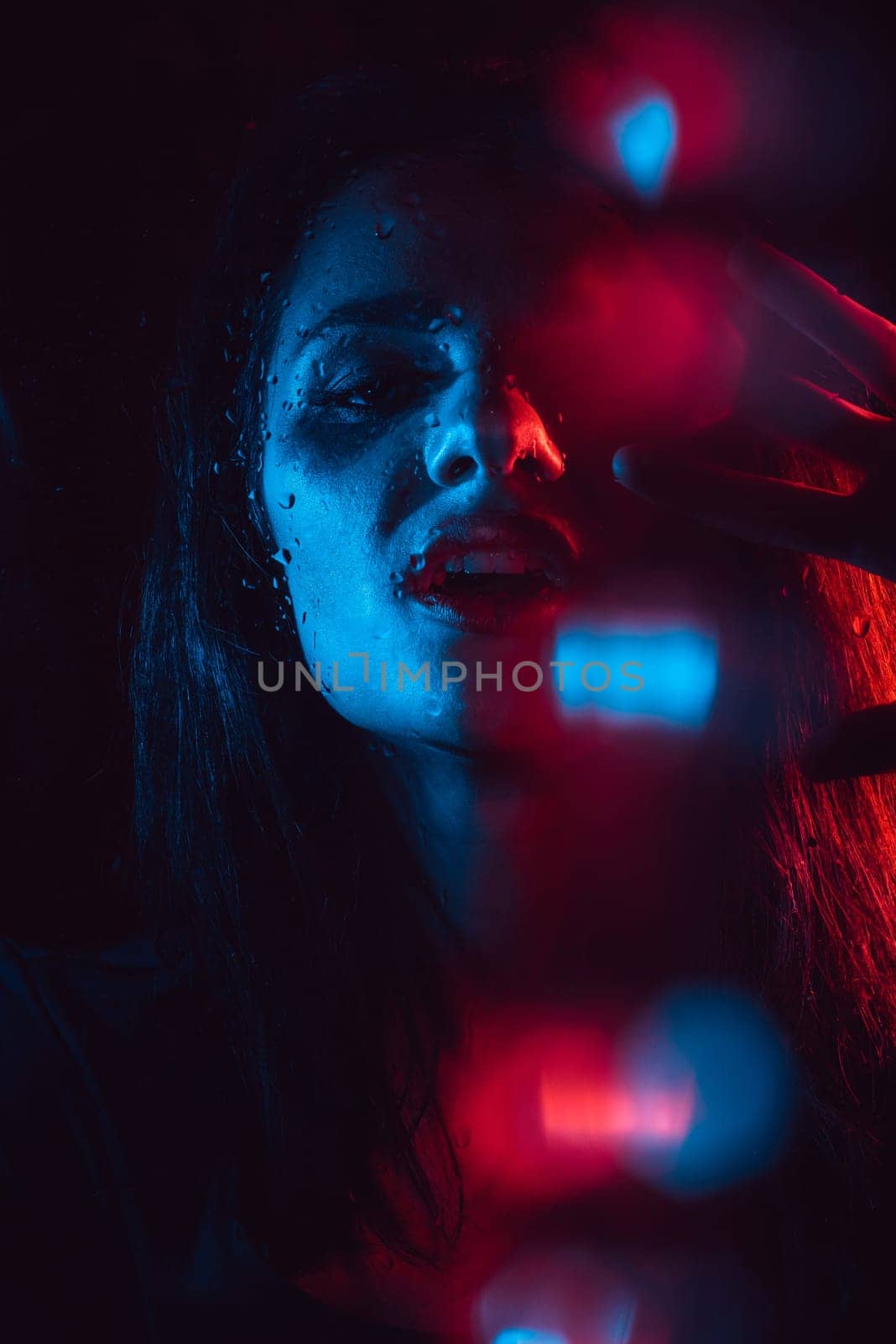 Sensual portrait of sad melancholic lonely girl behind glass with raindrops and neon red-blue lighting