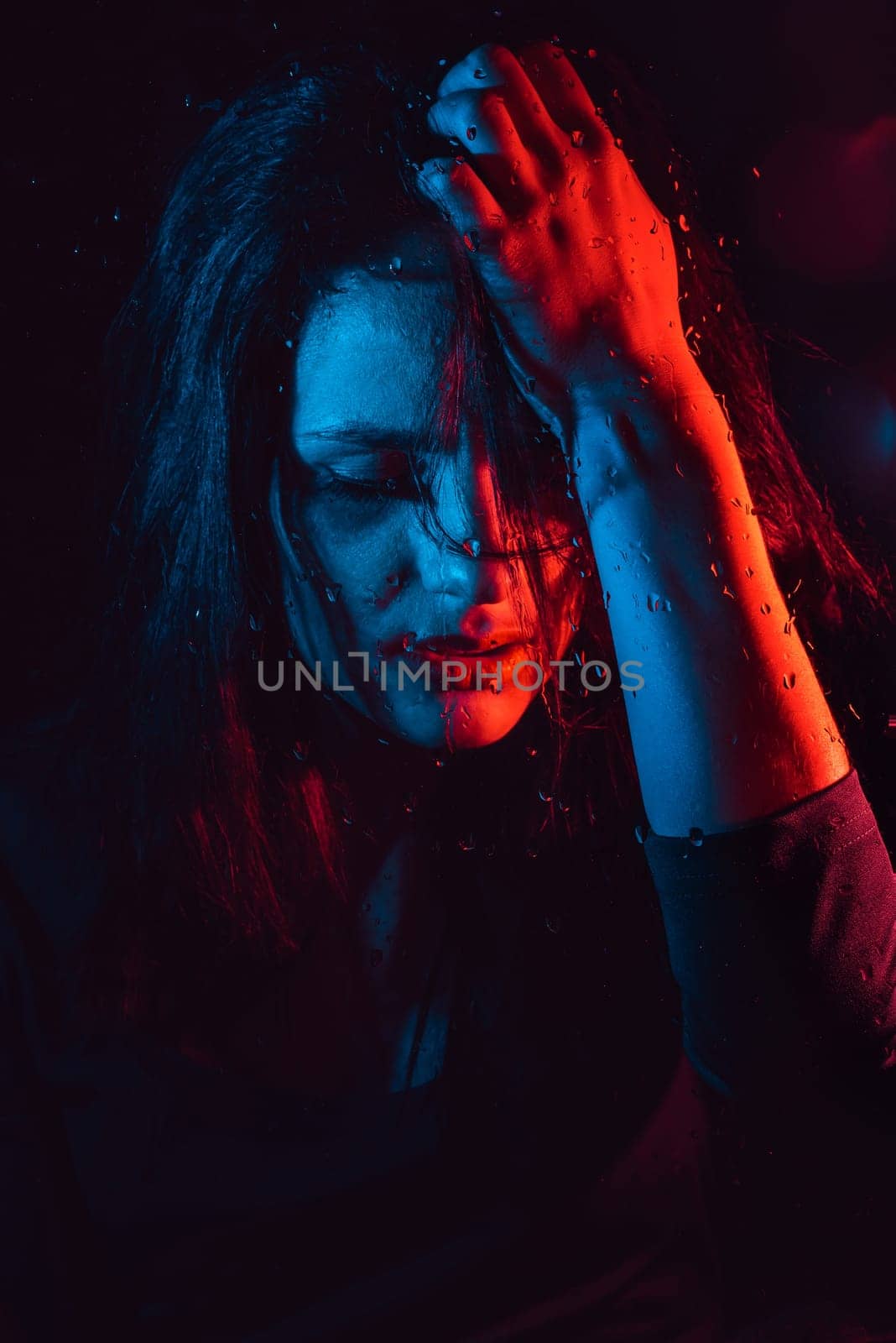 Sensual portrait of beautiful attractive brunette girl behind glass with raindrops with colored red blue neon lighting