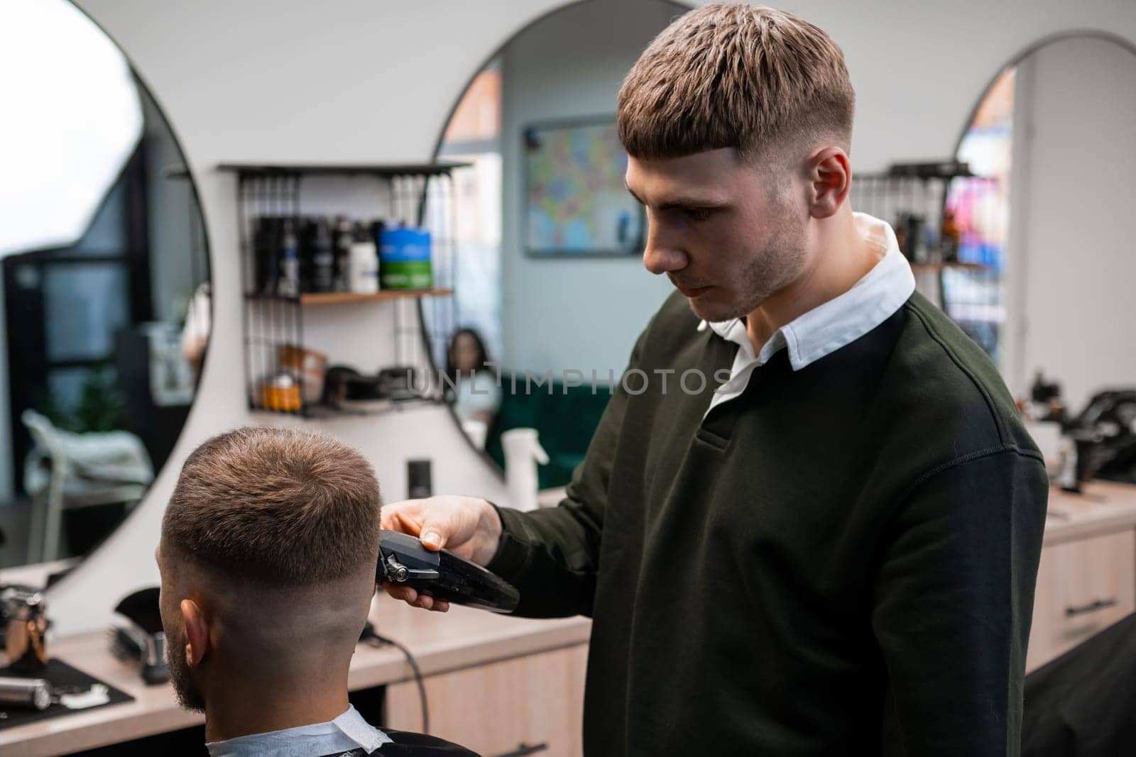 Barber uses shaver machine to cut young man hair in barbershop closeup. Master does modern haircut to male client in professional salon