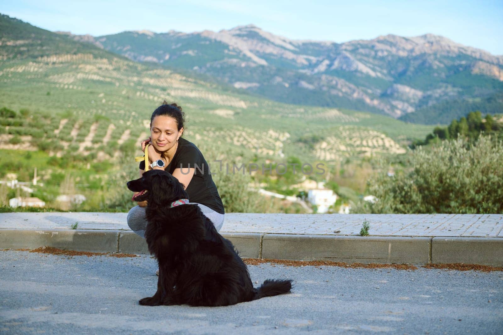 Smiling young woman sitting with her dog in the mountains, enjoying a happy time with her pedigree black cocker spaniel pet, dreamily looking aside into the distance. People and playing pets