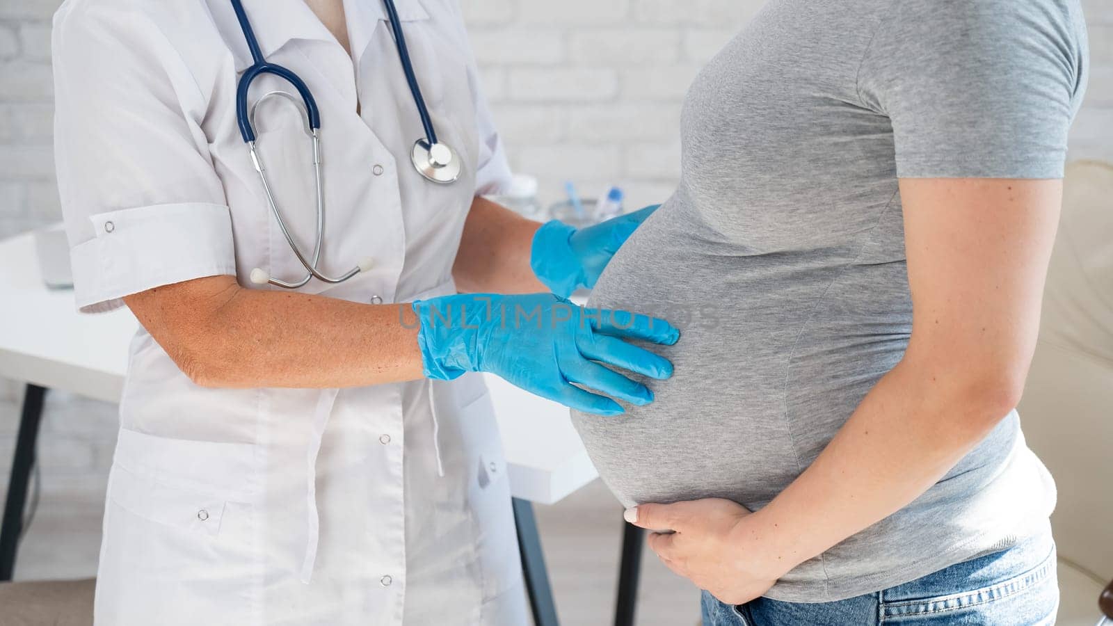 Pregnant woman visiting a doctor. Elderly Caucasian female gynecologist holds hands on the tummy of a pregnant patient