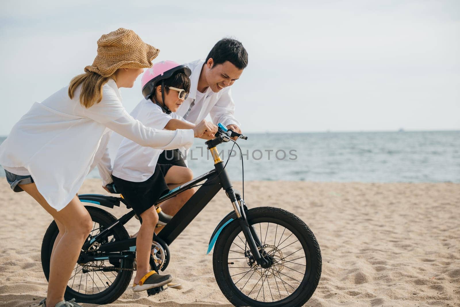 Happy family moments on summer road trip, Parents teach their children to ride bicycles on sandy beach near sea. Smiles safety helmets and carefree spirit of childhood fill this cheerful scene. by Sorapop