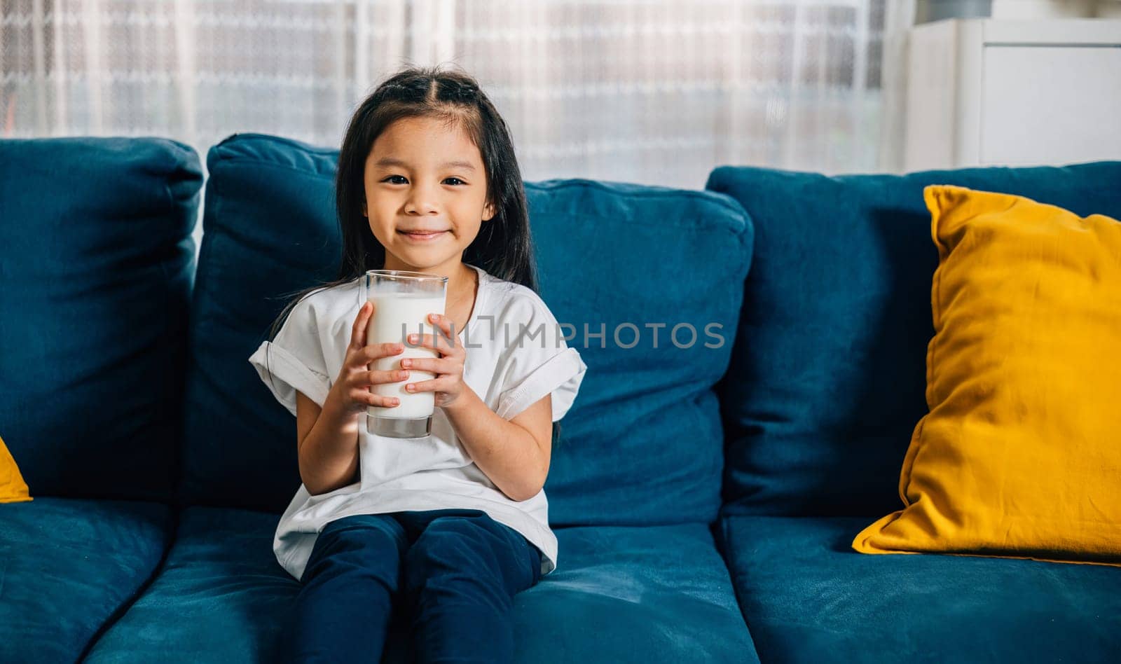 An adorable Asian child enjoys a glass of milk on the comfy sofa at home radiating happiness and innocence. This snapshot captures the essence of daily health care and nutrition in childhood.