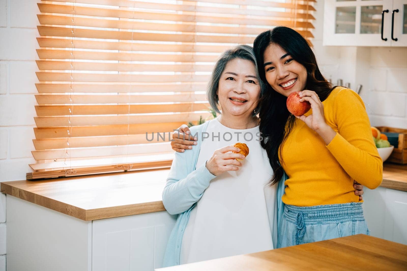 In the house, an old mother and her daughter, a young woman, stand together in the kitchen. They are holding an apple, showcasing the joy of learning and teaching, family, and togetherness. by Sorapop
