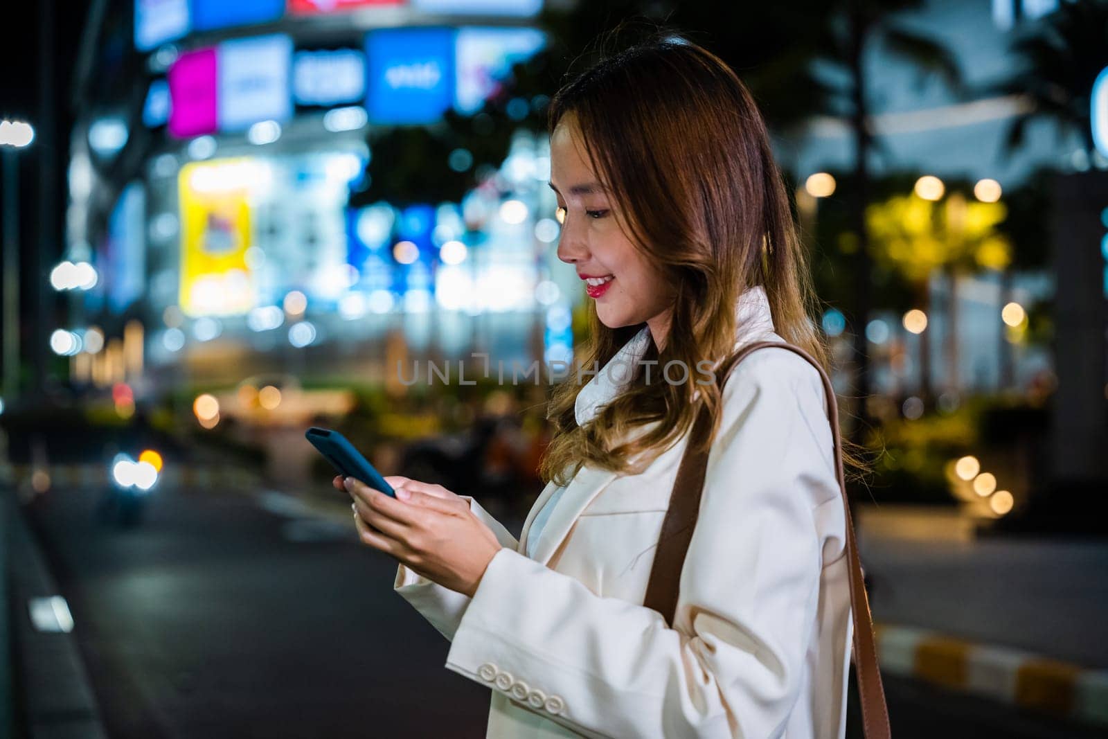 A portrait of a woman standing on the street at night, holding her smartphone and waiting for a ride.
