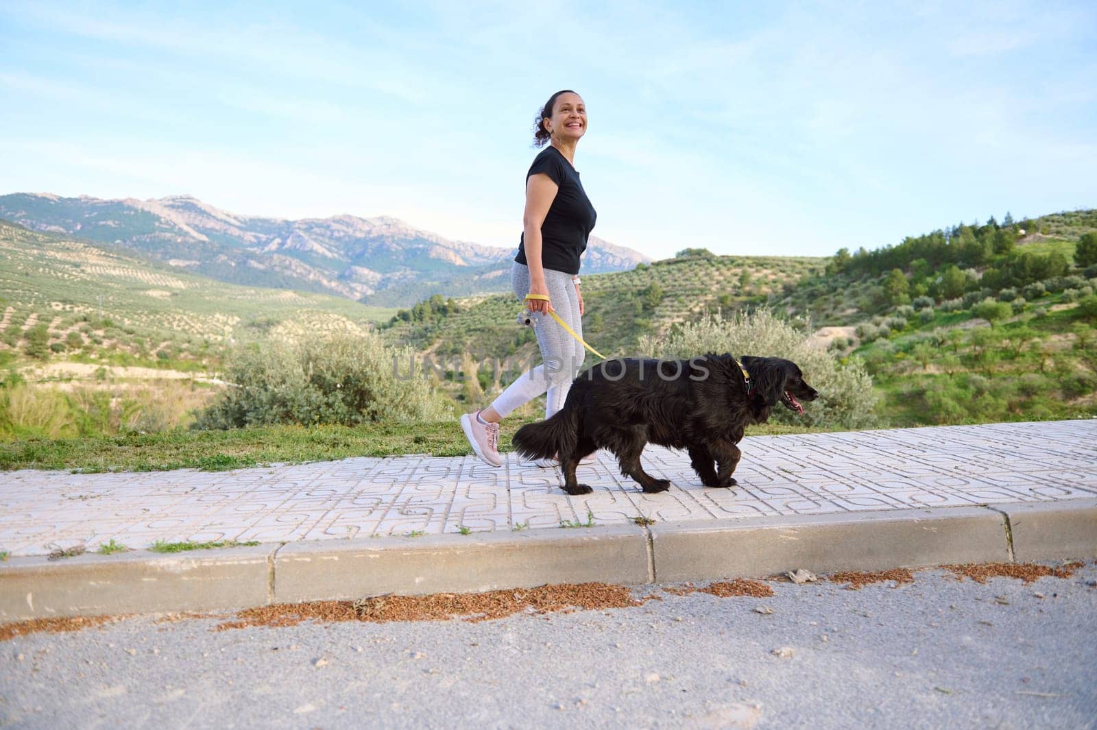Full length shot of a woman in active wear, walking her dog on the mountains nature. Domestic animals, pets and people concept