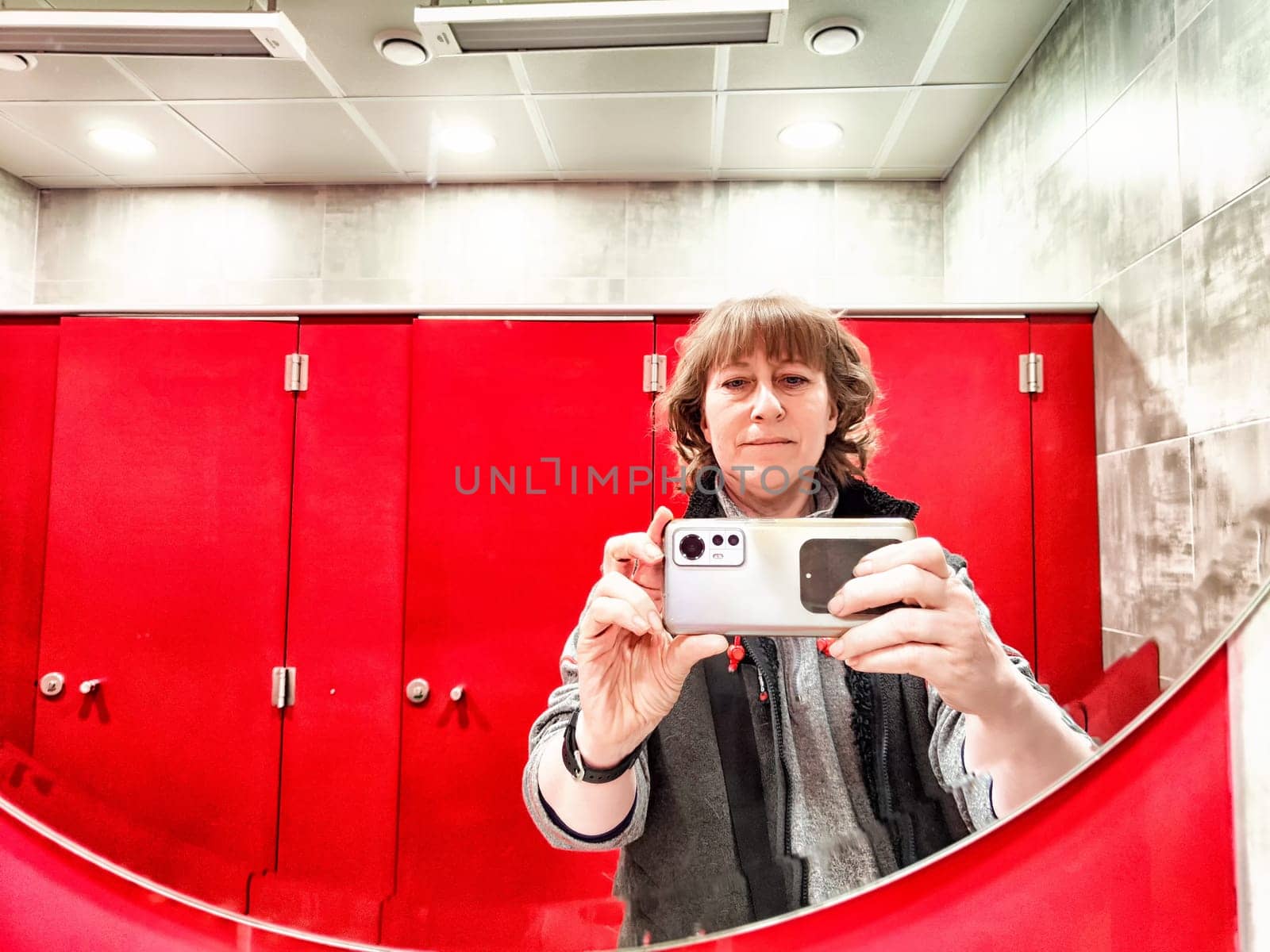 Middle-Aged Woman Taking a Selfie in Mirror With Red Lockers. A woman captures a selfie, mirrored against vibrant red lockers