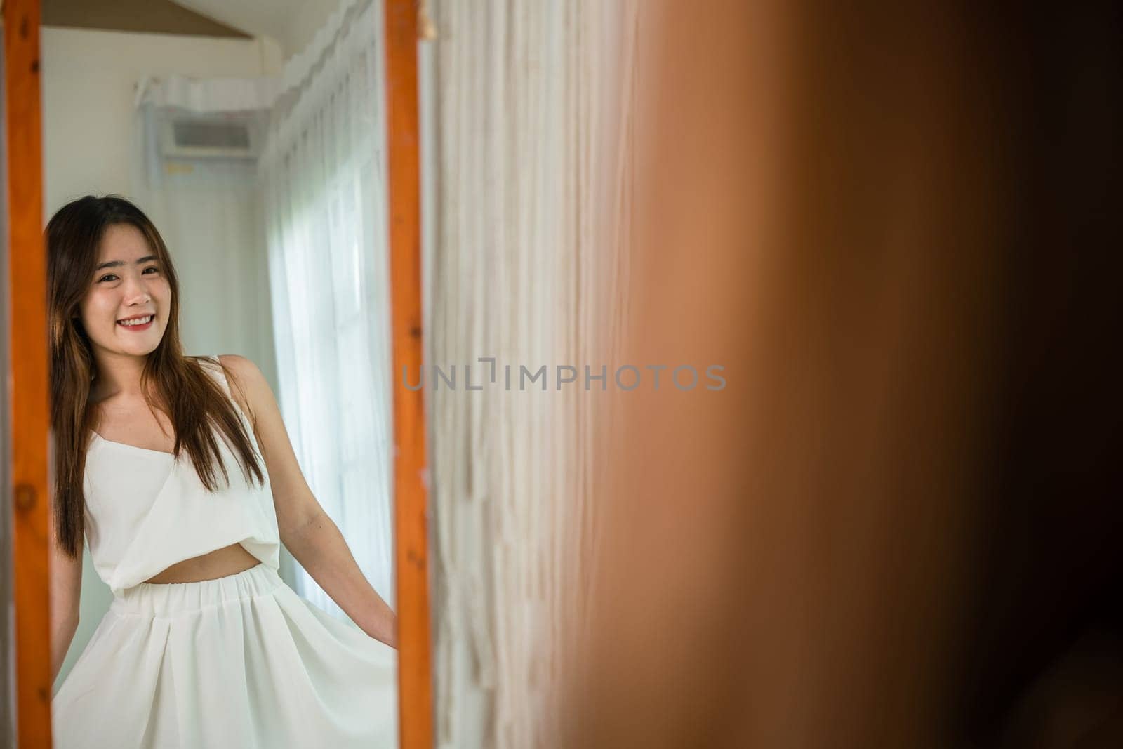 Asian beautiful young woman trying on blouse and smiling while standing in front of mirror in room, Happy female she wear on front mirror looking dress at home after morning wake up