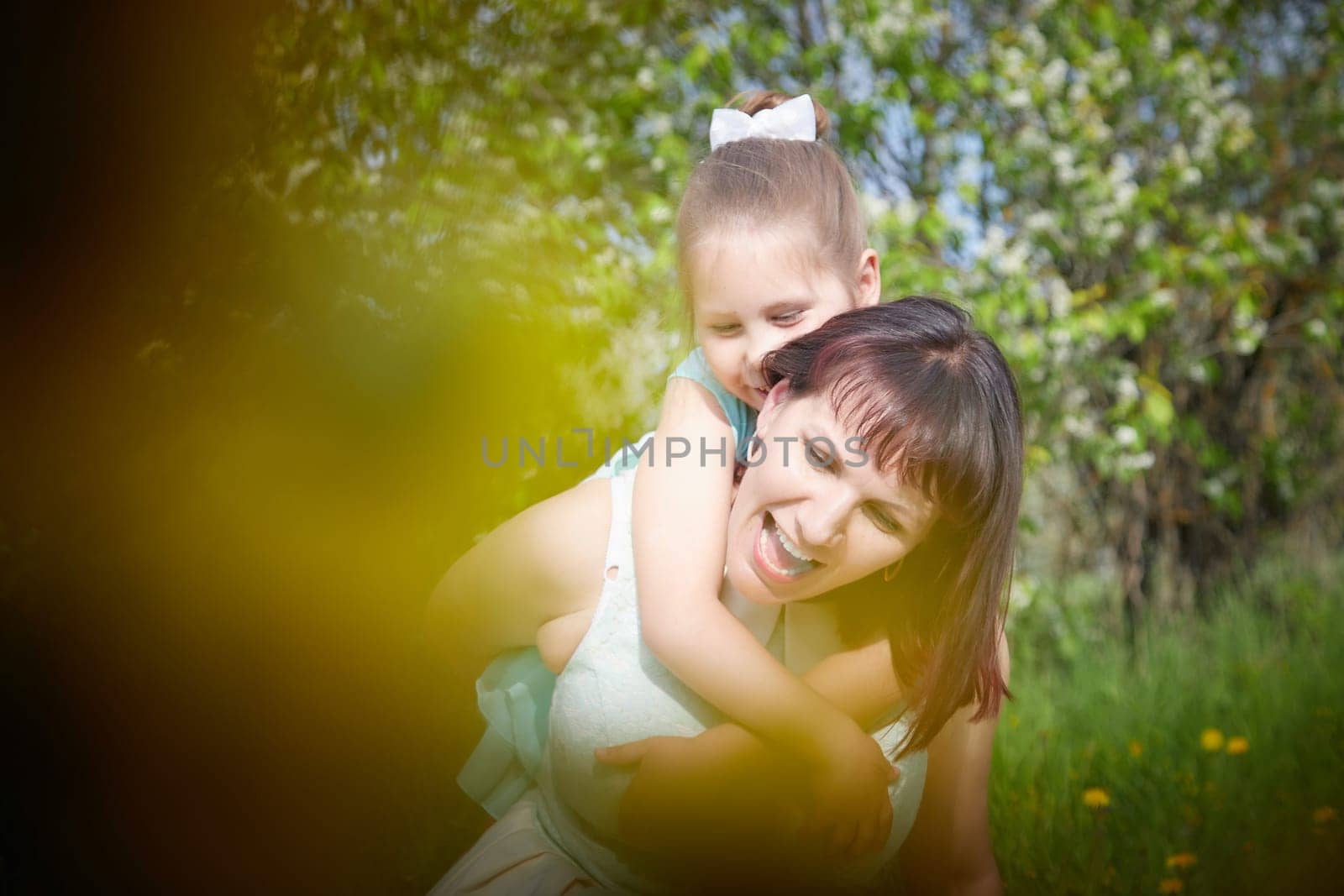 Happy mother and daughter enjoying rest, playing and fun on nature on a green lawn with dandelions and blooming apple tree on background. Woman and girl resting outdoors in summer and spring day