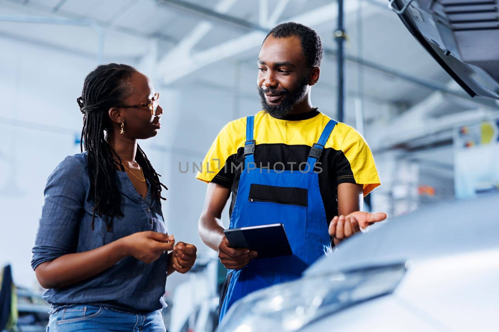 Happy mechanic in car service ordering new brakes for damaged vehicle using tablet. Smiling technician next to customer looking online for components to replace old ones in malfunctioning automobile