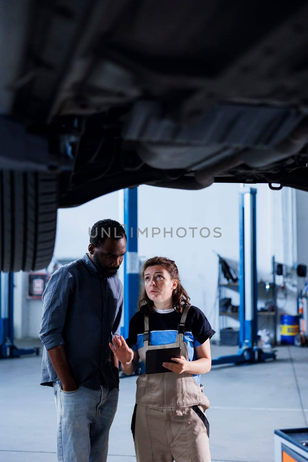 Knowledgeable mechanic in garage using laptop to follow checklist while doing maintenance on car, talking with client. Specialist in auto repair shop does checkup on vehicle tires next to customer