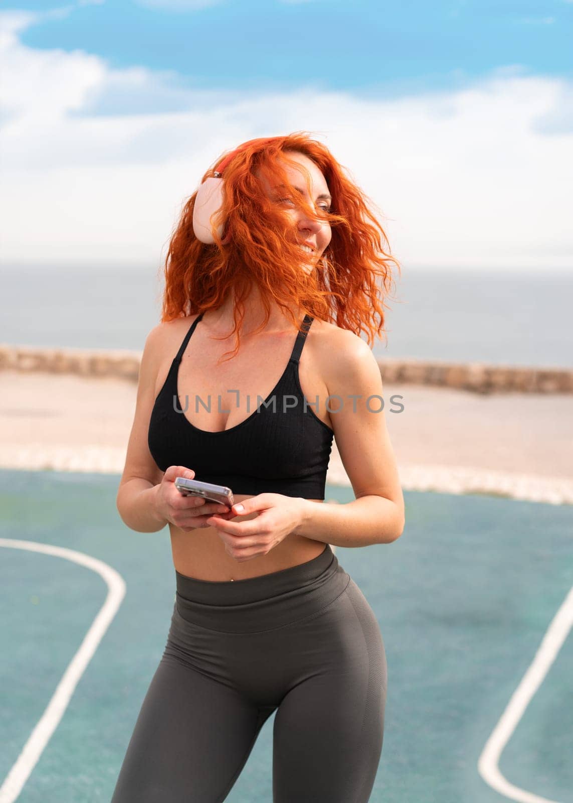 Happy redhead fit woman holding smartphone while listening to music through wireless headphones. Smiling female athlete in sportswear with swaying red hair standing at gym park on sunny day.