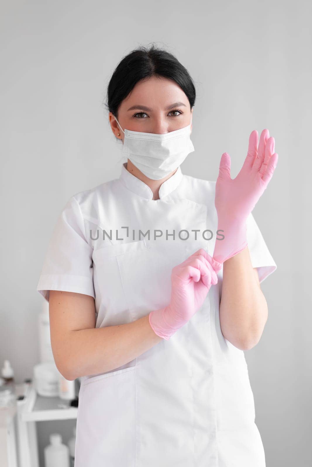 Female cosmetologist in mask putting on pink gloves before beauty treatment in clinic. Confident woman dermatologist in uniform working in hospital.