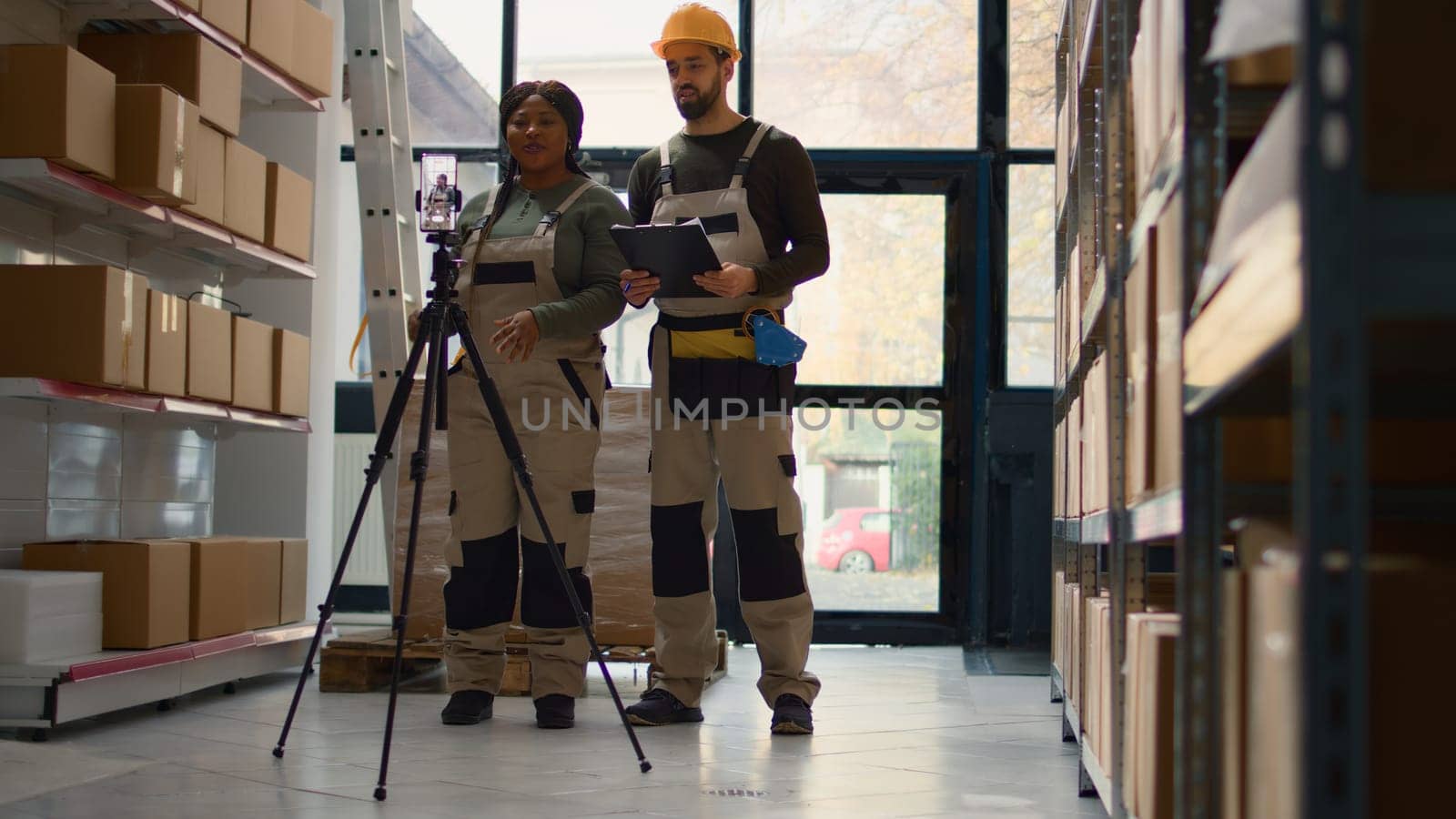 Warehouse head of operations and coworker using smartphone placed on tripod to make training video for interns. Directors film themselves in fulfillment center showing trainees how to store boxes