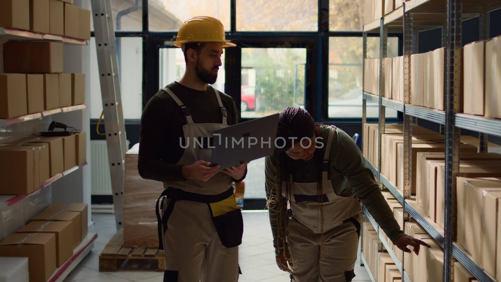 Employee and helpful colleague inputing updated products info on online shopping website, walking through warehouse storage center rows checking labels on cardboard boxes sitting on shelves