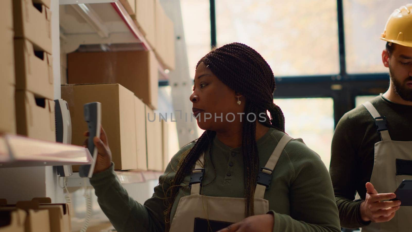 Logistics coordinator and warehouse picker preparing orders for delivery, receiving phone call in retail storehouse while scanning labels on cardboard packages to be shipped