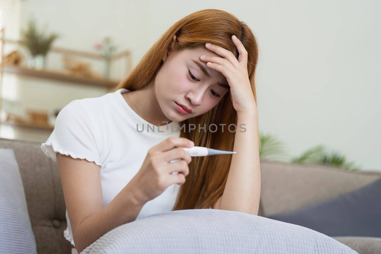 Sick young asian woman with thermometer suffering from cold on sofa, measuring body temperature with thermometer, touching forehead in sofa at home.