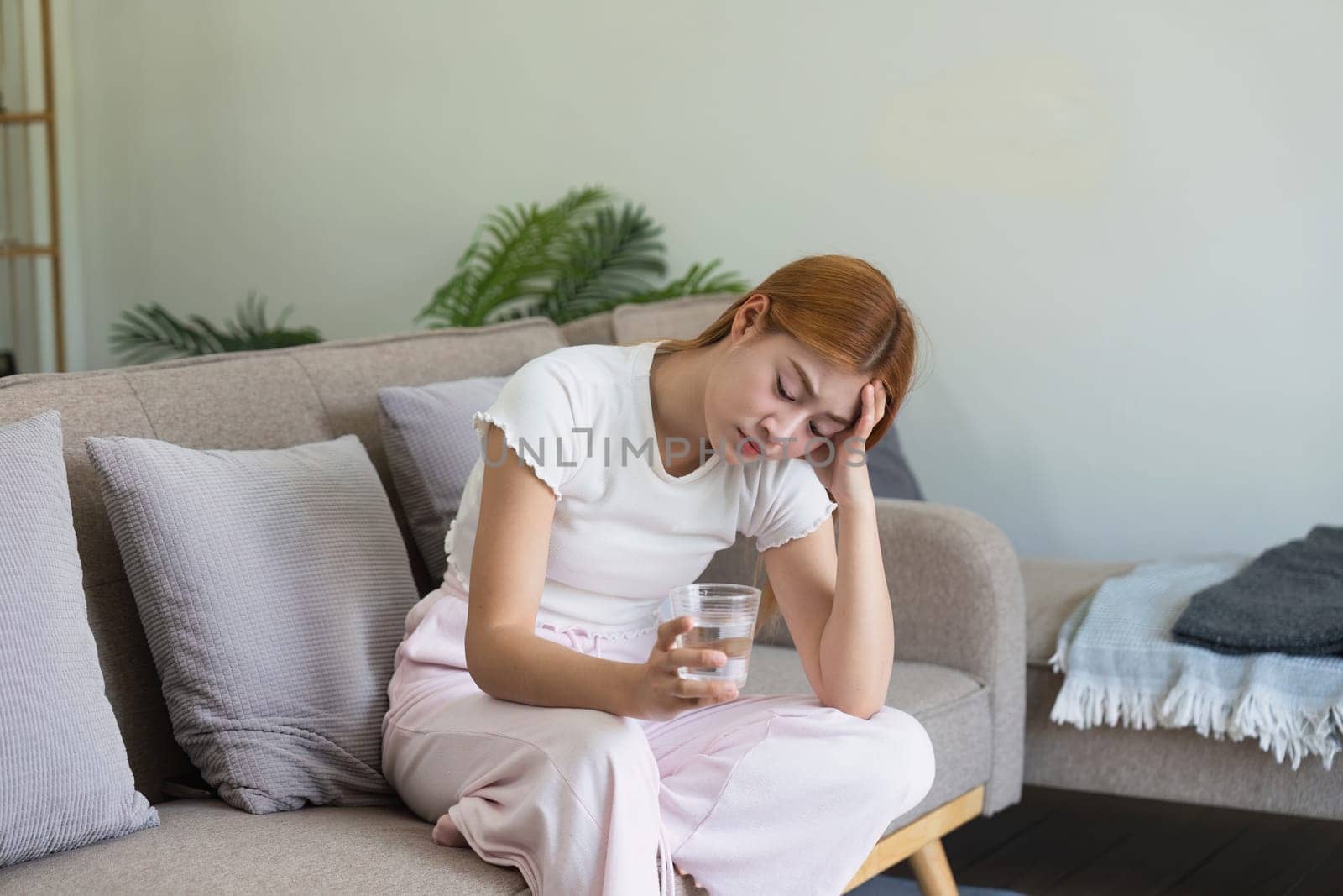 sick asian woman take medicine by water on sofa at home. having high fever while checking body temperature.