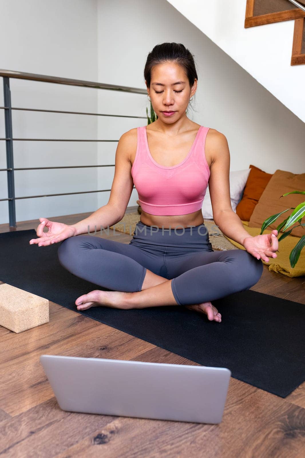 Young Asian woman meditating at home with online video meditation lesson using laptop. Vertical image. Healthy lifestyle concept.