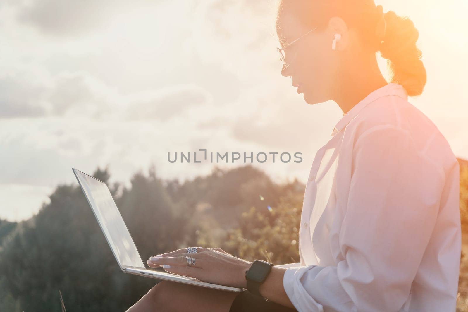 Digital nomad, woman in the hat, a business woman with a laptop sits on the rocks by the sea during sunset, makes a business transaction online from a distance. Freelance, remote work on vacation.