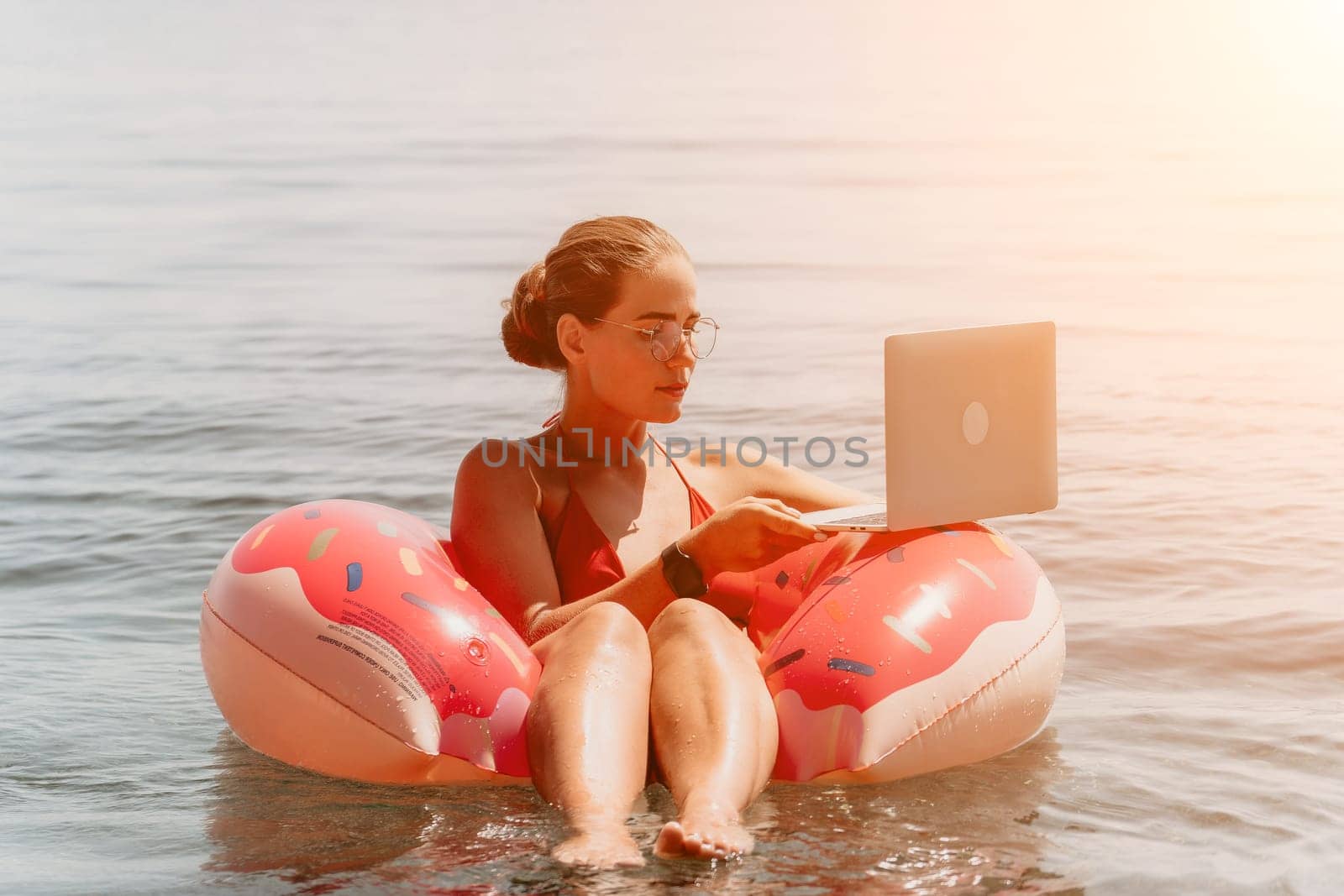 Woman freelancer works on laptop swimming in sea on pink inflatable ring. Pretty lady typing on computer while floating in the sea on inflatable donut at sunset. Freelance, remote work on vacation