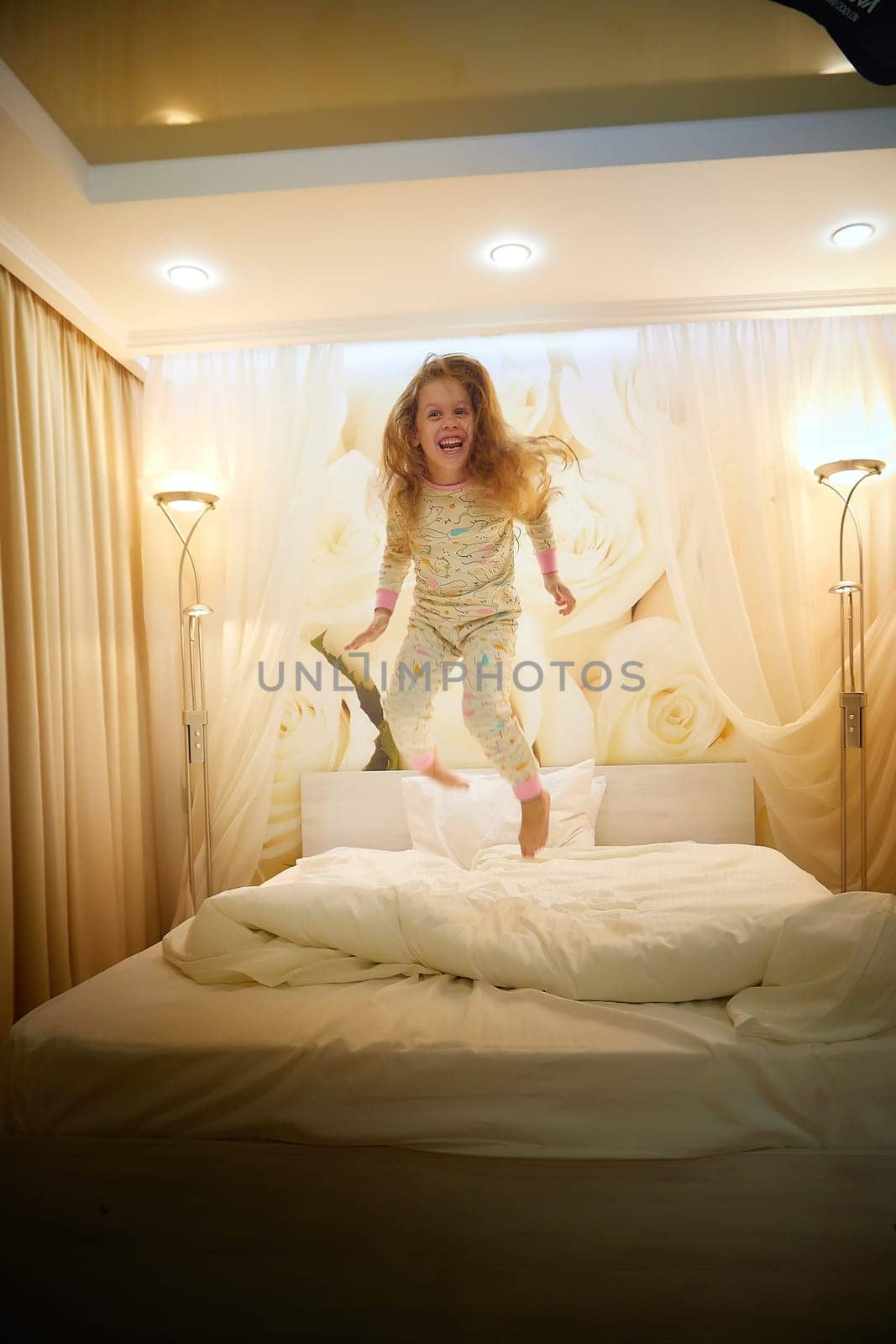 Cute little girl jumping on white bed in sleeping room or in hotel in evening