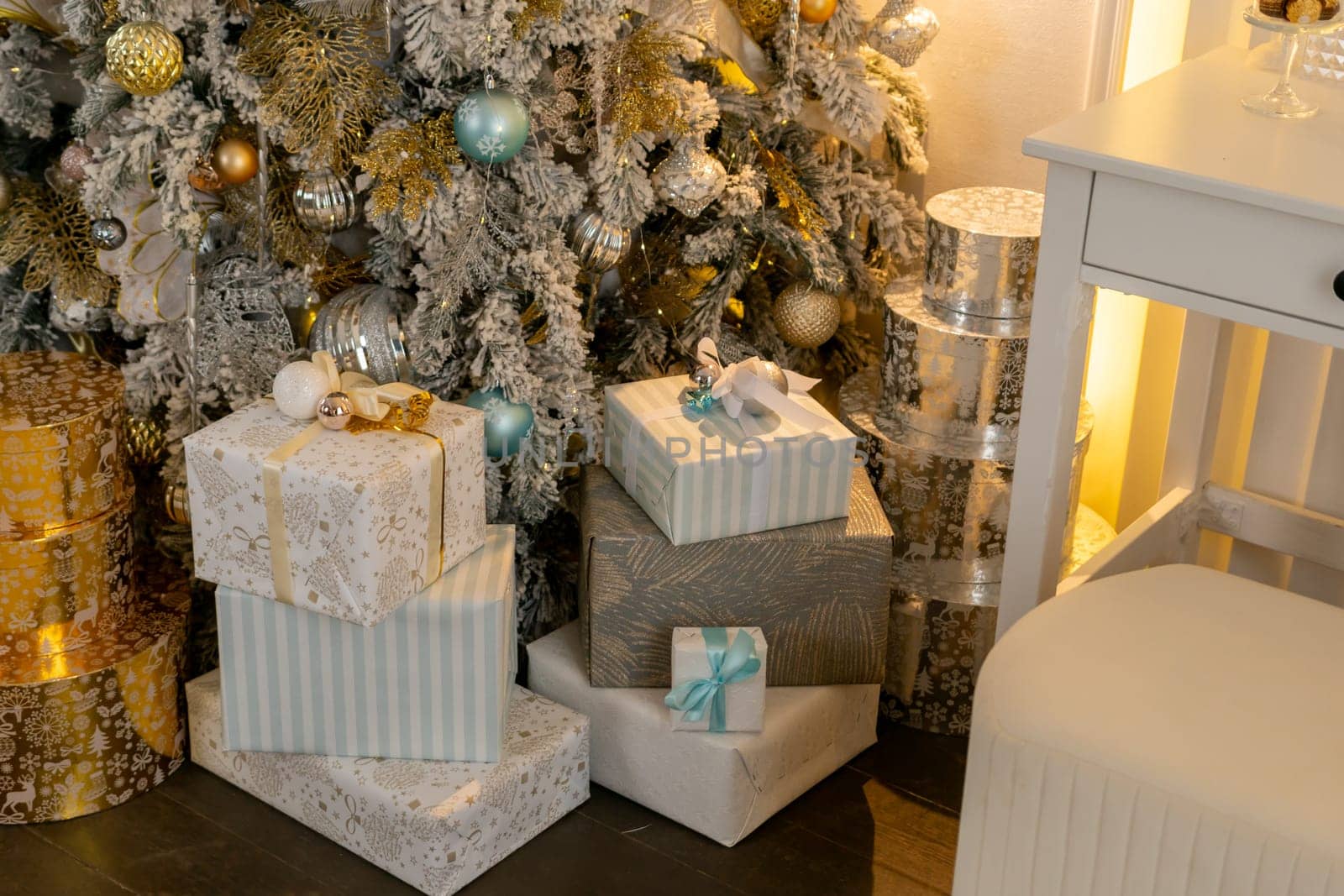 A Christmas tree with many gifts stacked on top of it. The gifts are wrapped in white paper and blue and gold ribbons