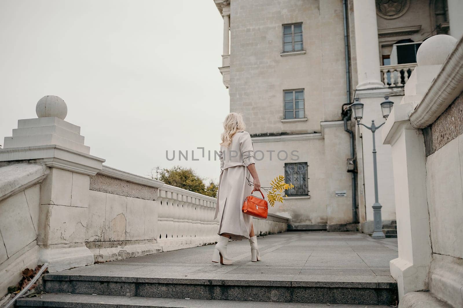 A woman wearing a white coat and carrying a red purse walks down a stone staircase. The scene is set in a city, with a large building in the background. The woman is in a hurry. by Matiunina