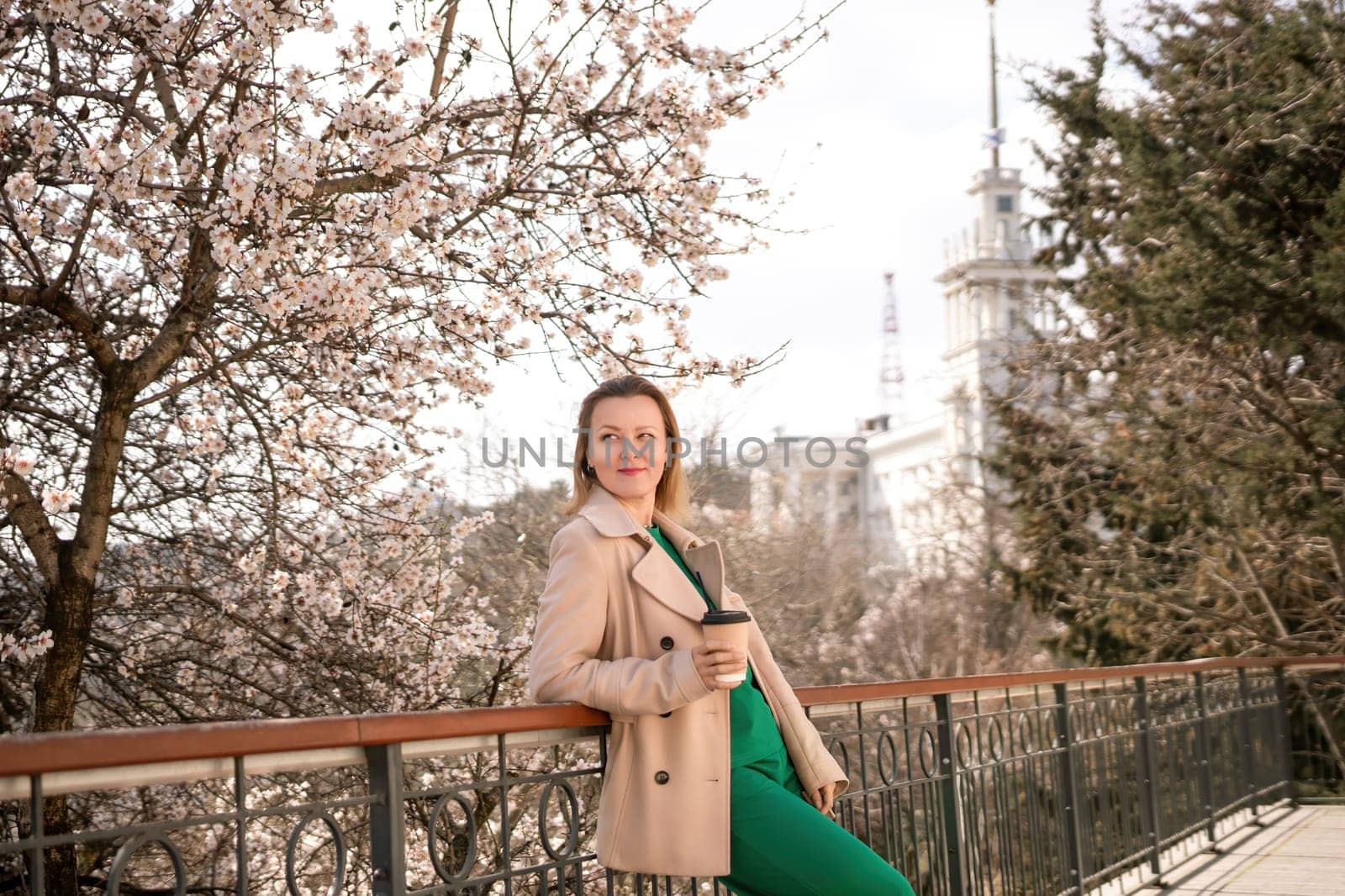 A woman is standing on a ledge with a cup of coffee in her hand. She is wearing sunglasses and a tan coat. The scene is set in a park with cherry blossoms in bloom. The woman is enjoying the view. by Matiunina
