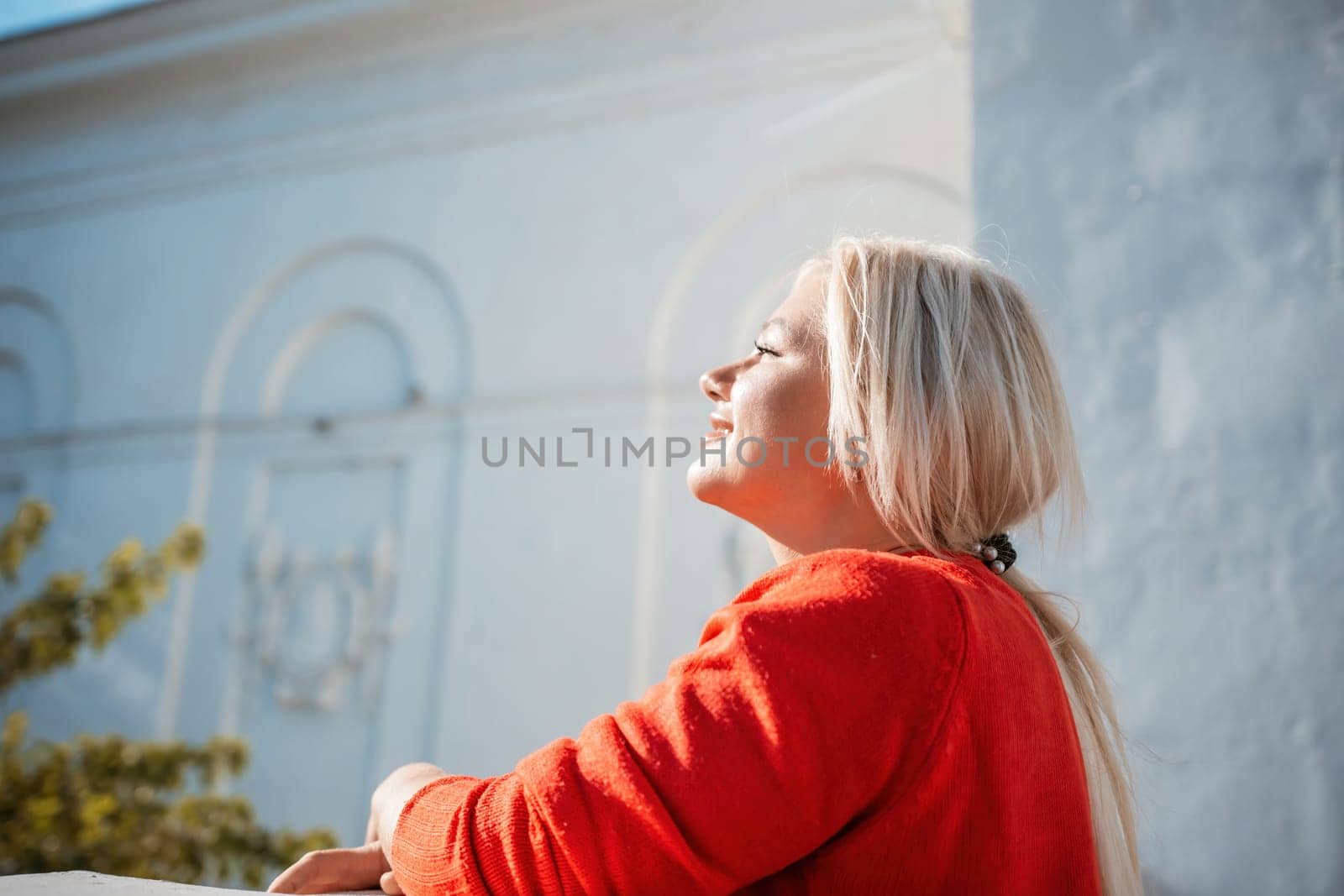 A woman in a red sweater is sitting on a ledge. She is smiling and looking out at the street. by Matiunina