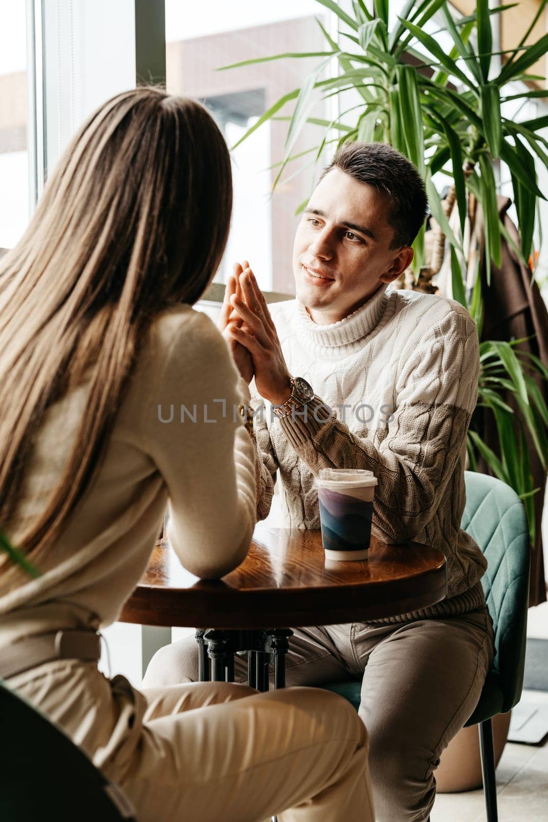A man and woman are seated opposite each other at a small table, deeply engaged in a conversation. The man, wearing a white knit sweater, listens attentively to the woman with his hands gently clasped. The warm ambiance of the cafe, with soft natural light spilling in, adds to the intimate setting.