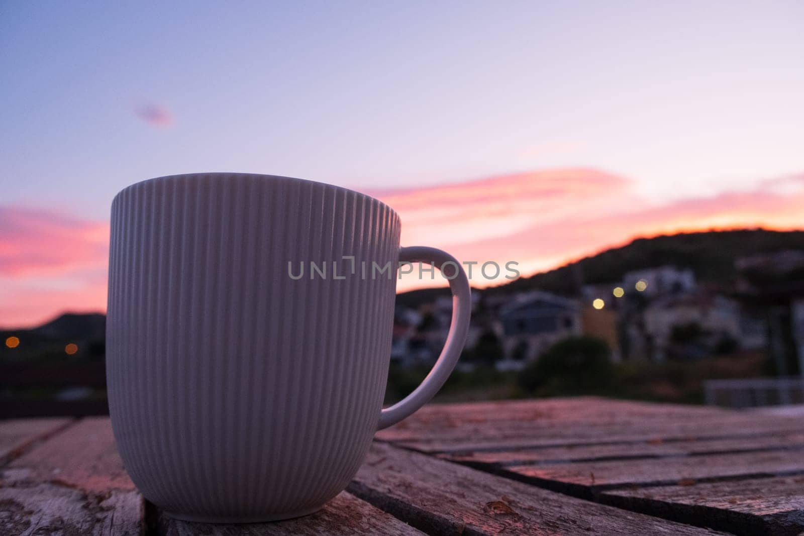As the sun sets, a coffee cup is placed on a wooden table against a backdrop of the colorful sky. The serene landscape is enhanced by the simple beauty of the tableware