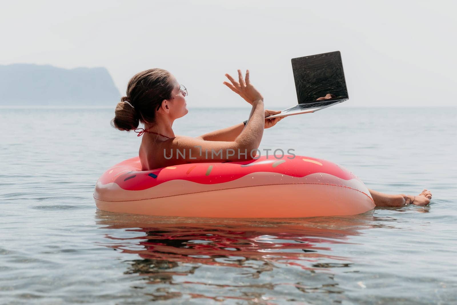 Woman freelancer works on laptop swimming in sea on pink inflatable ring. Pretty lady typing on computer while floating in the sea on inflatable donut at sunset. Freelance, remote work on vacation