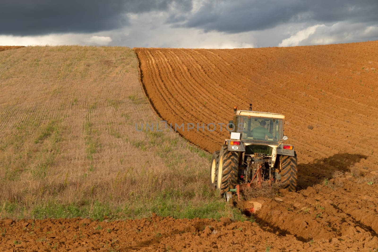 A tractor actively plowing a curved field with a stormy sky overhead, showcasing the dramatic contrast of agriculture and nature. by JavierdelCanto