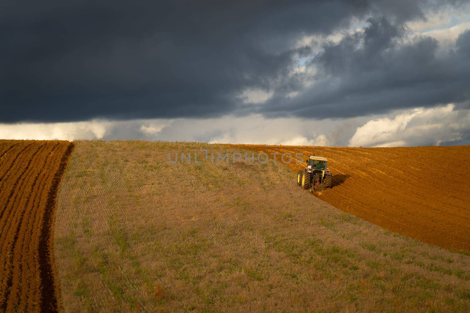 A tractor actively plowing a curved field with a stormy sky overhead, showcasing the dramatic contrast of agriculture and nature.